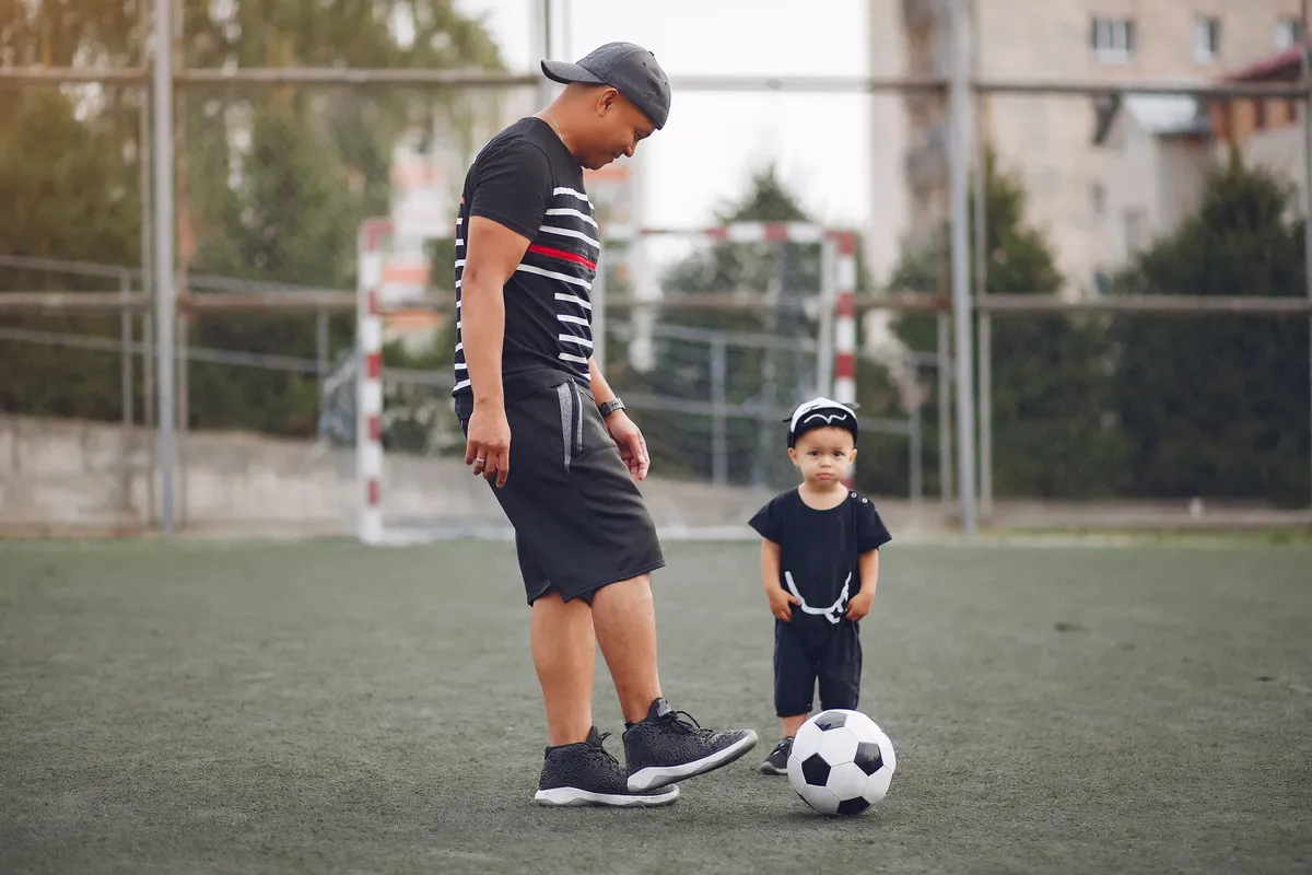 スポーツ場でのエチニティックな父が黒いカジュアルな服を着て、かわいらしい乳幼児の息子がフットボールを蹴る