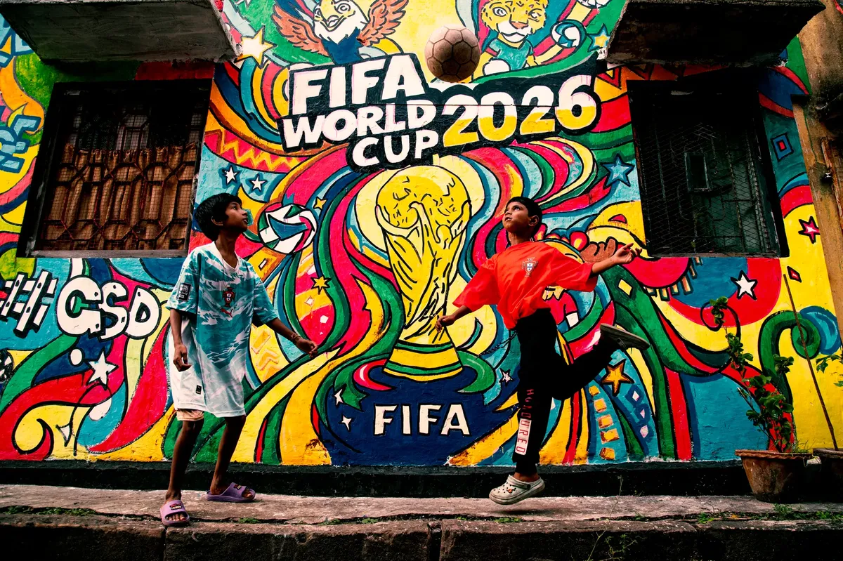 FIFAワールドカップ2026のエネルギッシュな壁画で、前景に子供たちがサッカーをしている様子が描かれています。