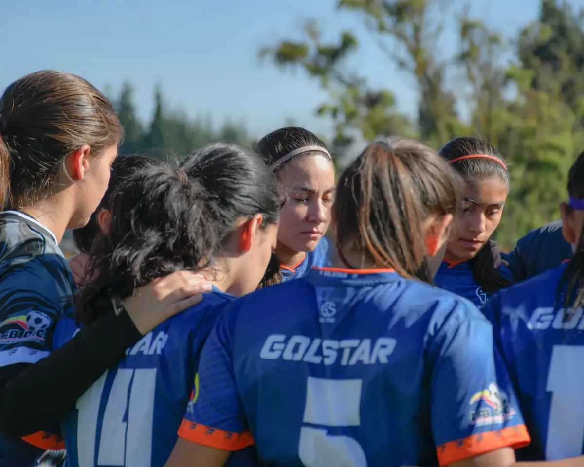 A women's soccer team unites in a pre-game huddle on a sunny day.