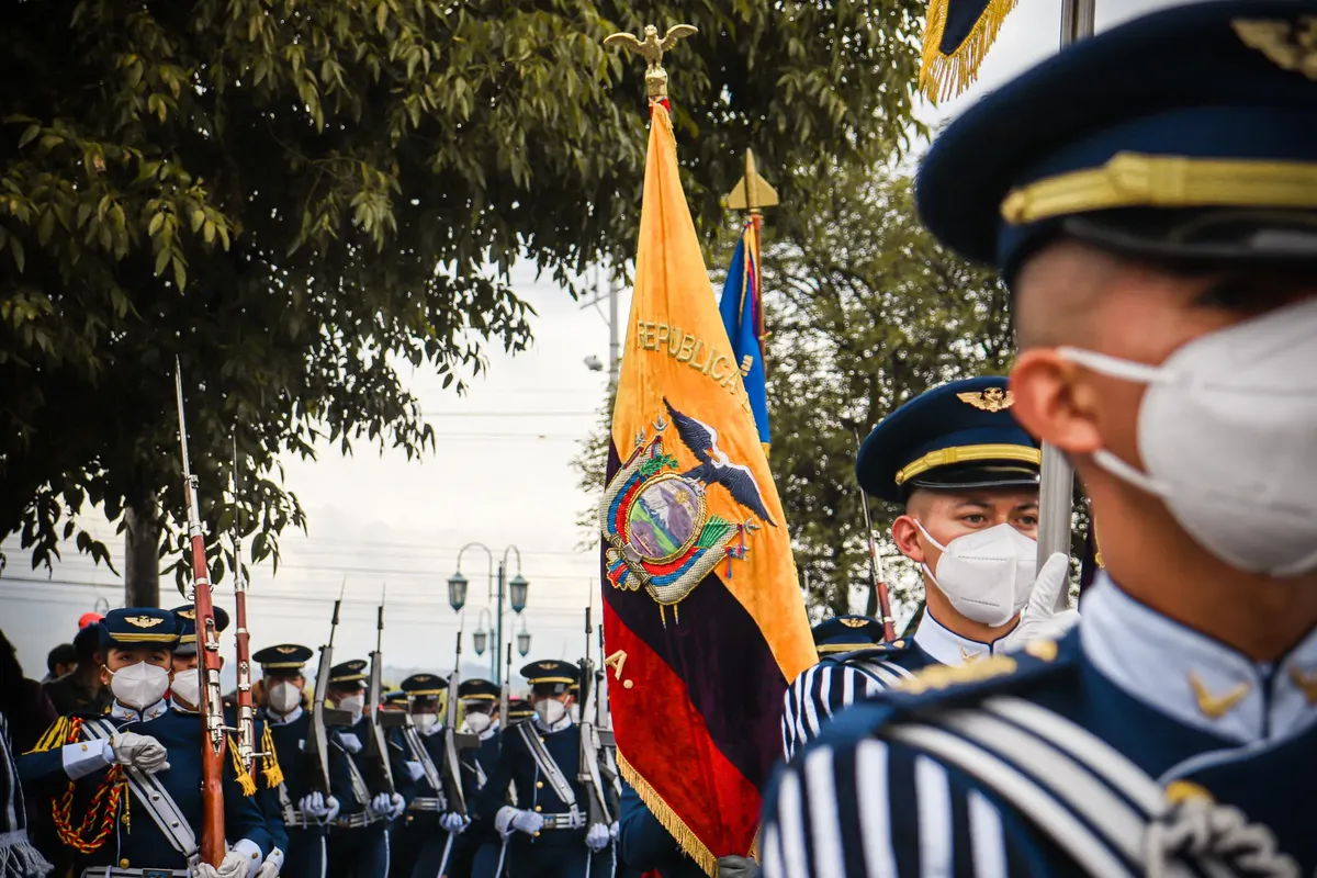 Military personnel in uniforms with masks march in an Ecuadorian parade featuring the national flag.